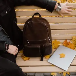 A person sitting on a bench with a backpack and laptop, surrounded by autumn leaves.